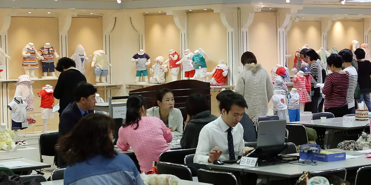 Employees working at long tables in a Korean company's headquarters, with product samples on shelves behind them